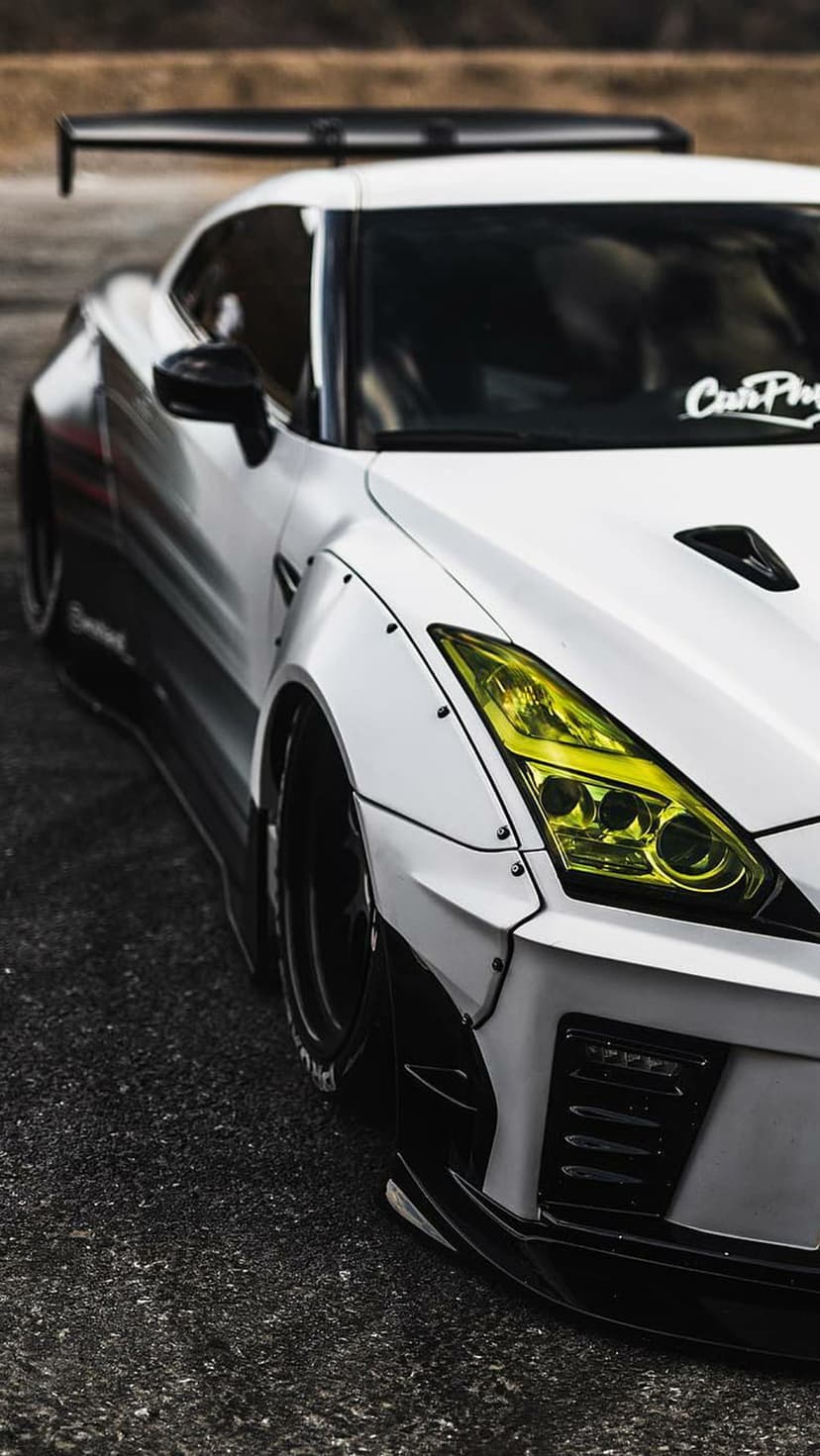 White Nissan GT-R with a widebody kit, yellow headlights, and a large rear wing parked on a dark asphalt street.