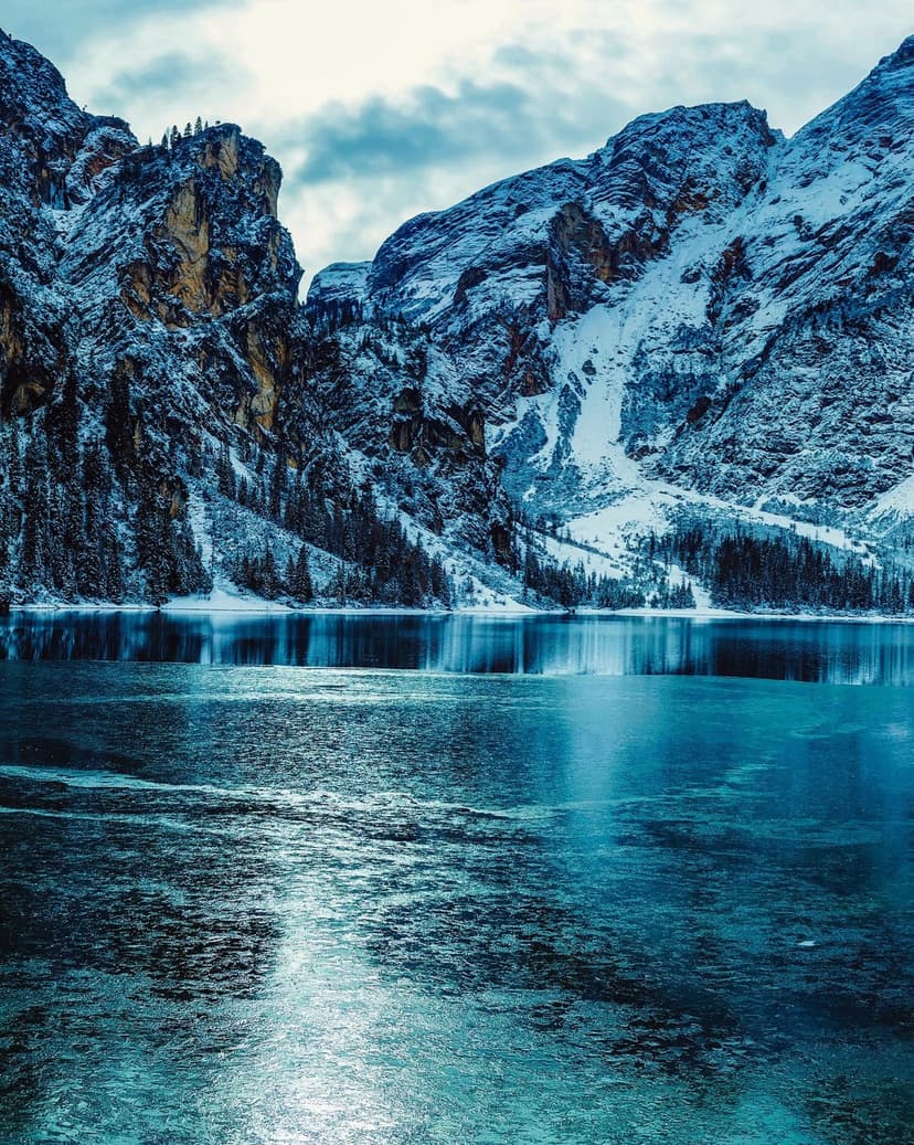 Snow-capped mountain peaks reflect in the calm, partially frozen waters of a deep blue alpine lake under clouds.