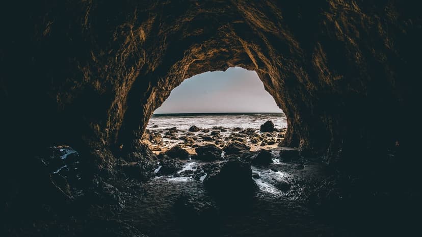 Craggy sea cave entrance frames a sunny ocean vista with foamy waves breaking against a jagged rocky coastline.