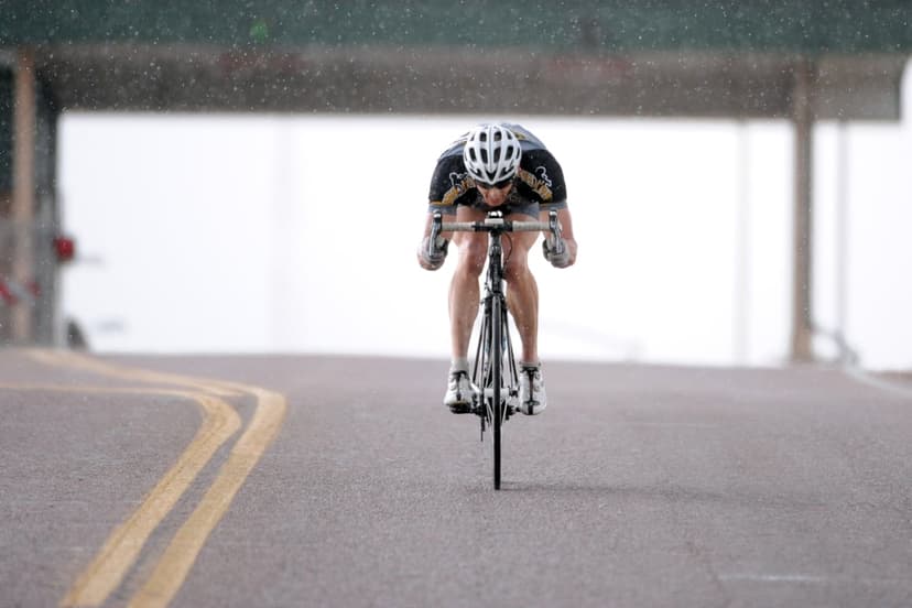 Cyclist in an aerodynamic tuck races down a wet asphalt road with yellow lines under falling rain and mist.