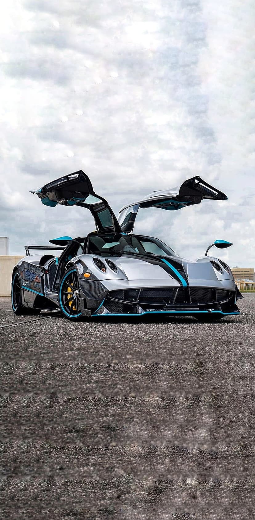 Silver Pagani Huayra hypercar with blue accents and open butterfly doors parked on textured asphalt outdoors.