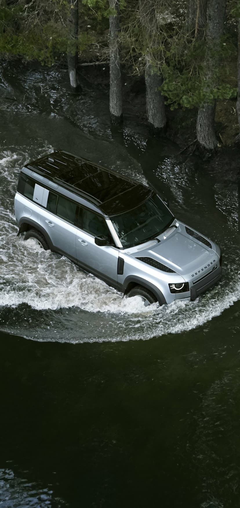Silver Land Rover Defender driving through a dark river, creating white water splashes in a forest setting.