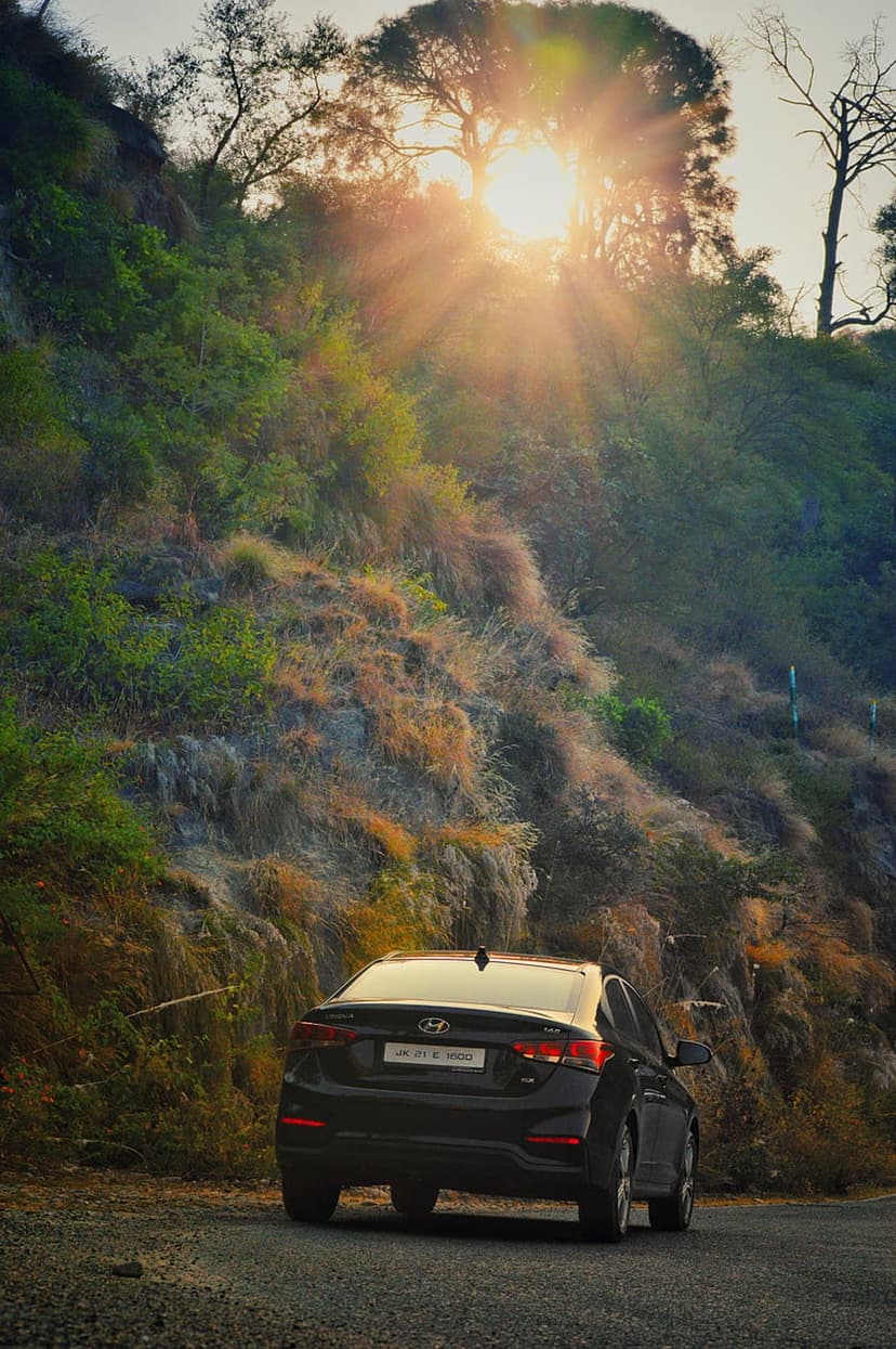 Black Hyundai Verna sedan on a gravel mountain road with golden sunlight streaming through dense evergreen trees.