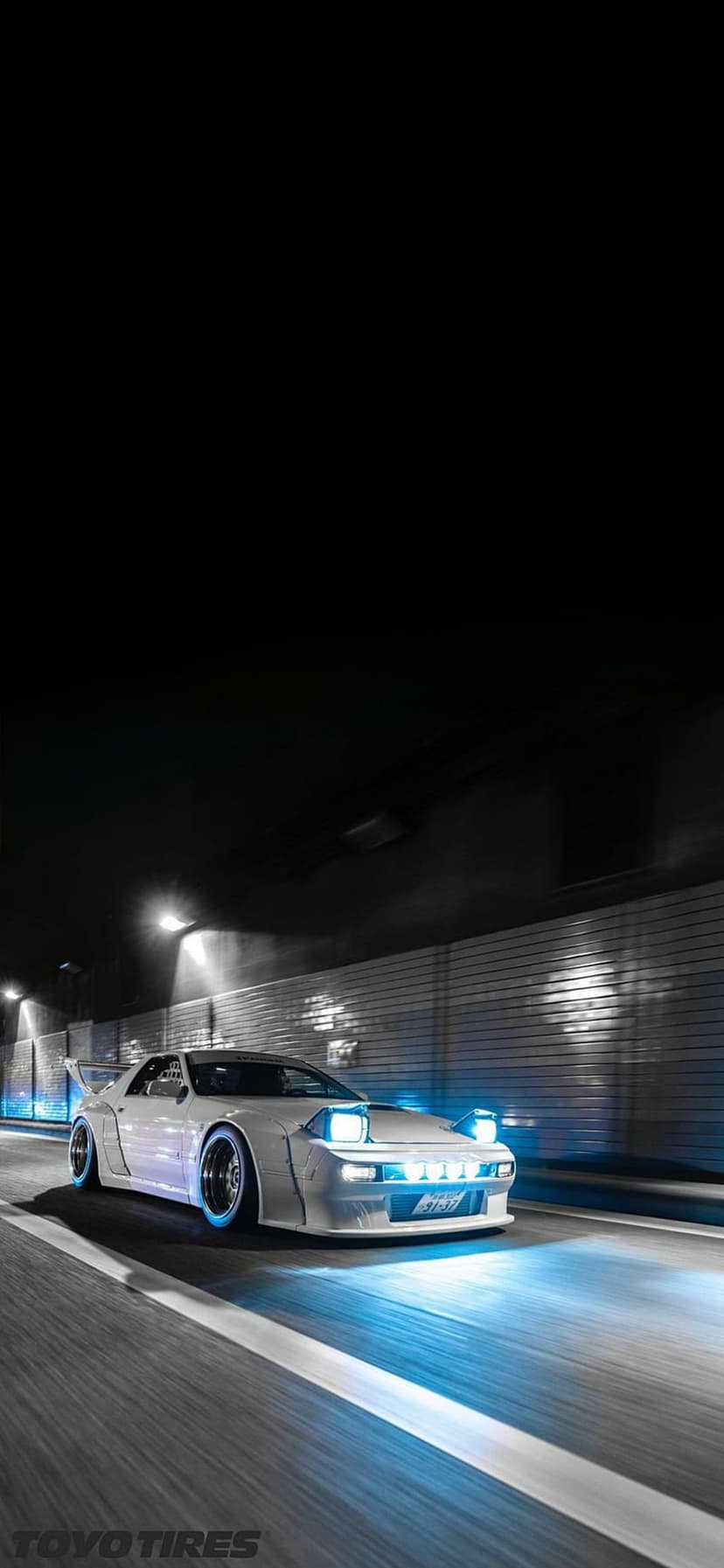 White sports car with glowing pop-up headlights and blue neon underglow speeding on a dark, wet urban road.
