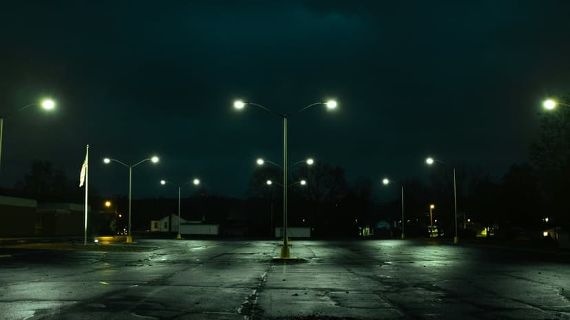 Empty asphalt parking lot at night with glowing streetlights casting long shadows on wet, cracked pavement.
