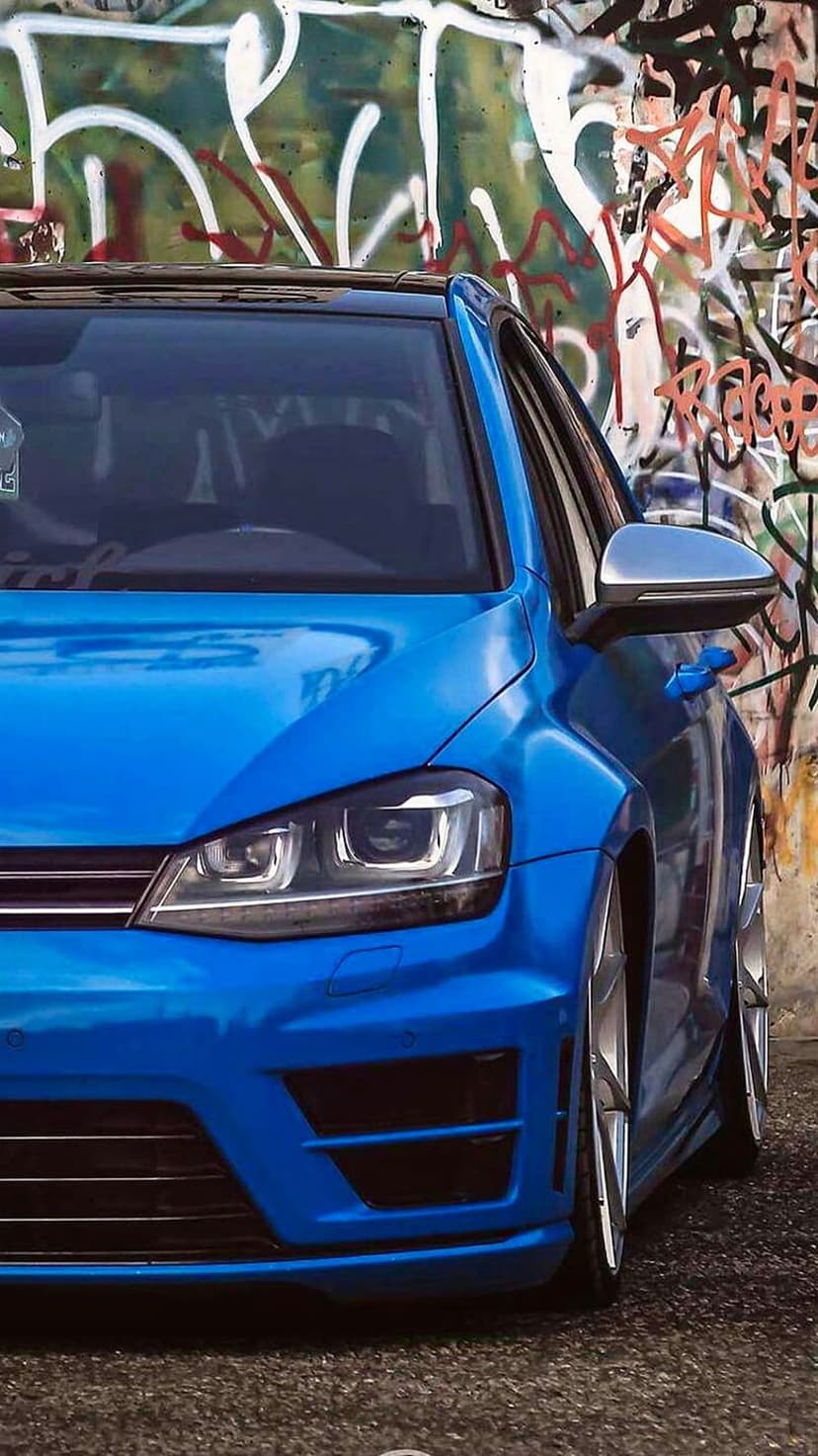 Blue sports car with custom wheels parked in front of a colorful graffiti mural on a textured brick wall.