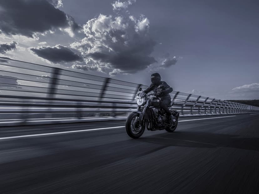 Black motorcycle speeding on a bridge with motion blur against a dramatic cloudy sky in desaturated tones.