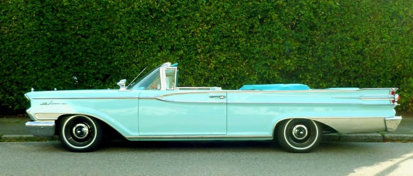Turquoise vintage convertible car with chrome accents parked on asphalt against a dense green garden hedge.