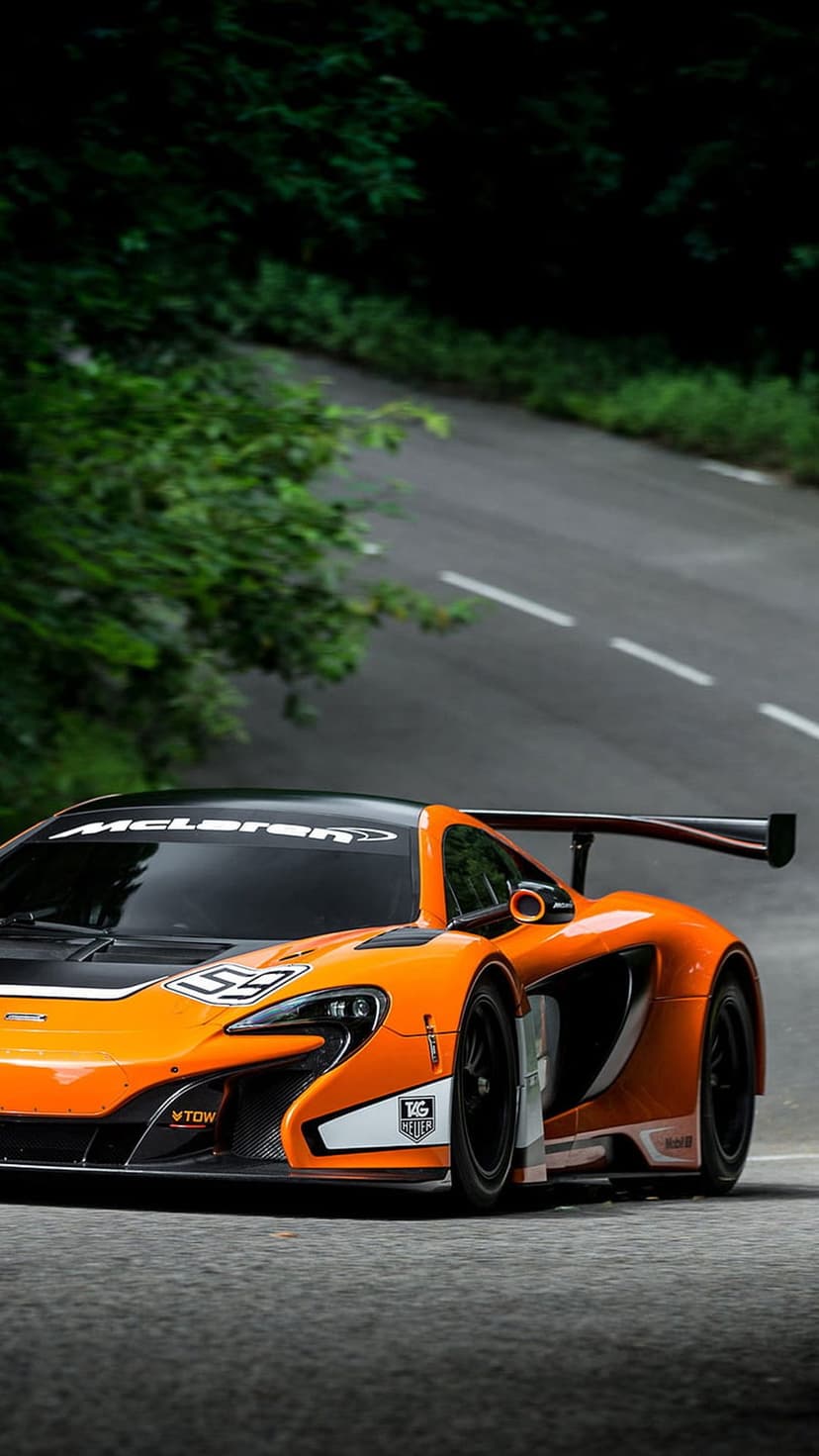 Orange McLaren race car with a large rear wing drives on a winding asphalt road surrounded by green forest trees.