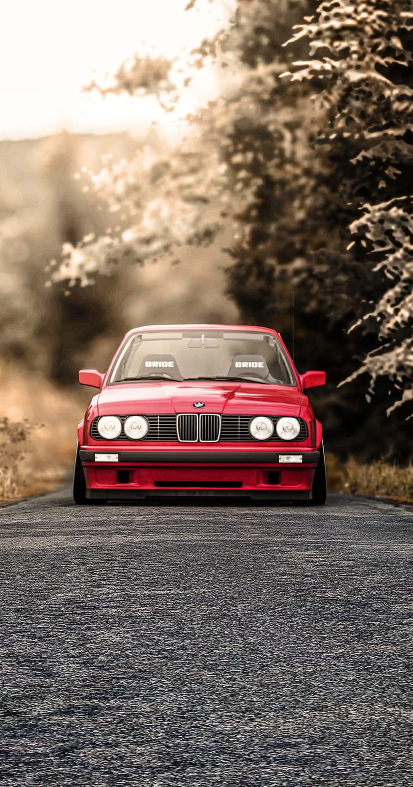 Red BMW E30 with Bride racing seats parked on asphalt with sunlight filtering through a blurred forest background.