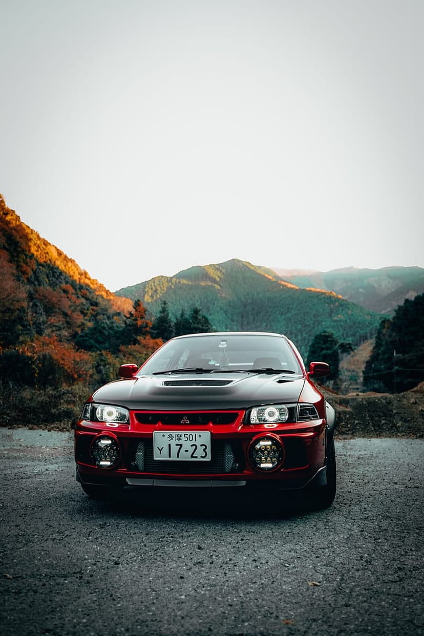 Red Mitsubishi Lancer Evolution with custom headlights parked on a gravel road near orange autumn mountain forests.