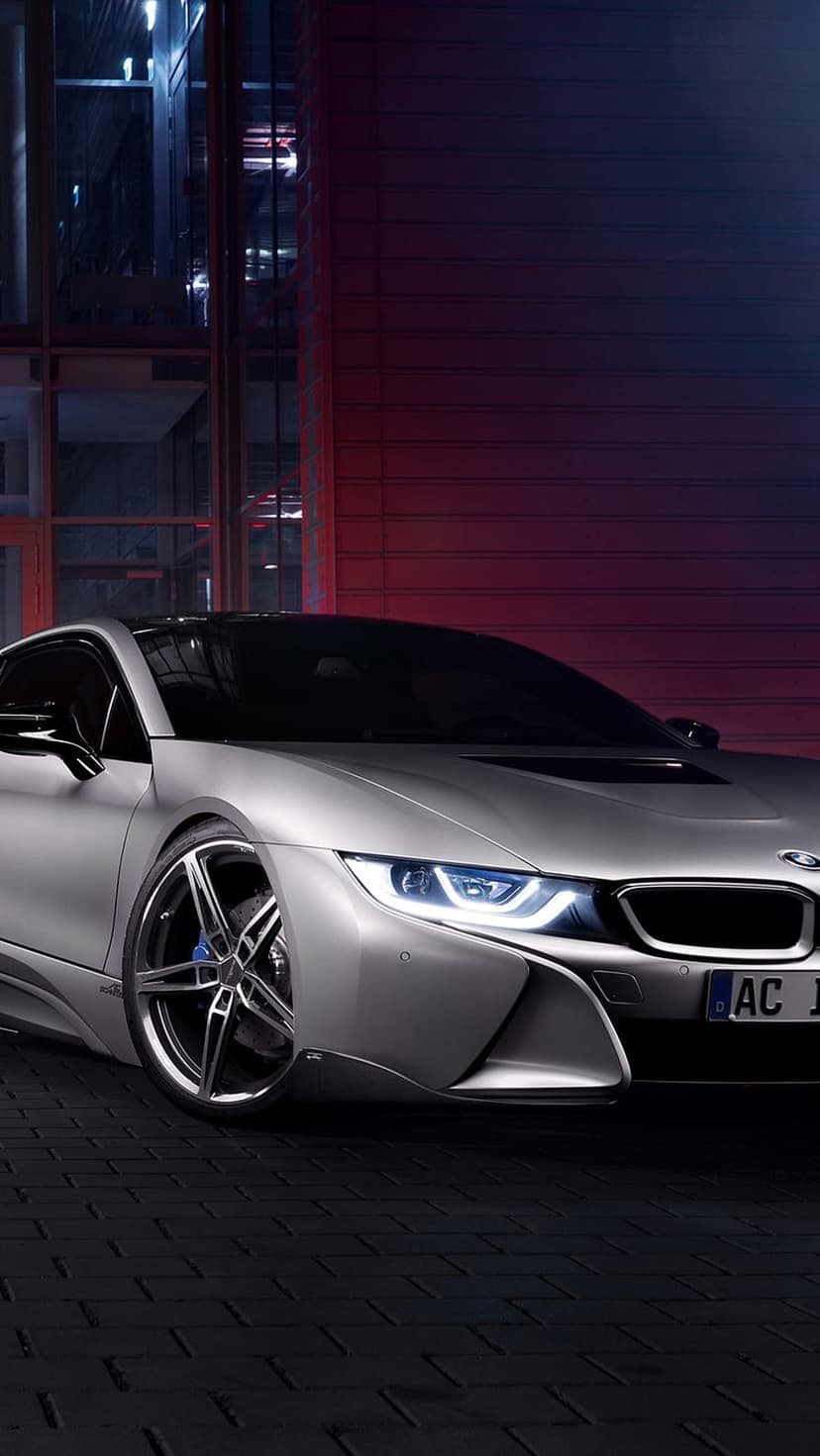 Silver BMW i8 sports car with blue glowing headlights parked on a brick surface near a red wall at night.