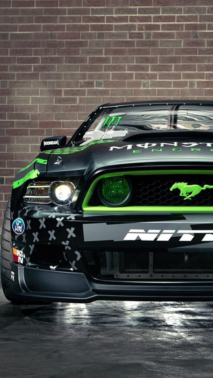 Custom Ford Mustang front grille with green Monster Energy branding and glowing headlights by a brick wall.