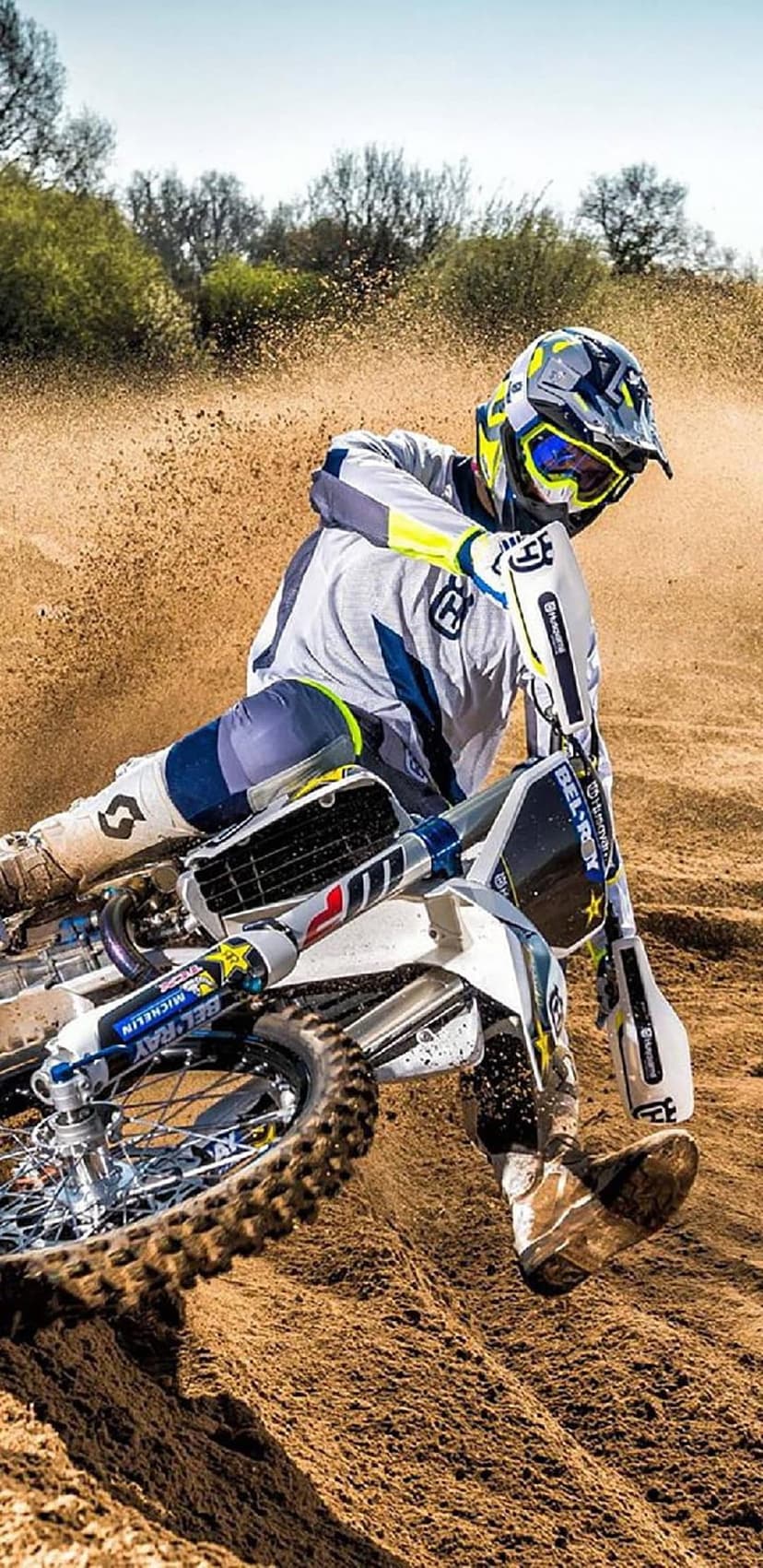 Motocross rider in yellow goggles leans into a dirt track turn, kicking up a dust cloud under a clear blue sky.