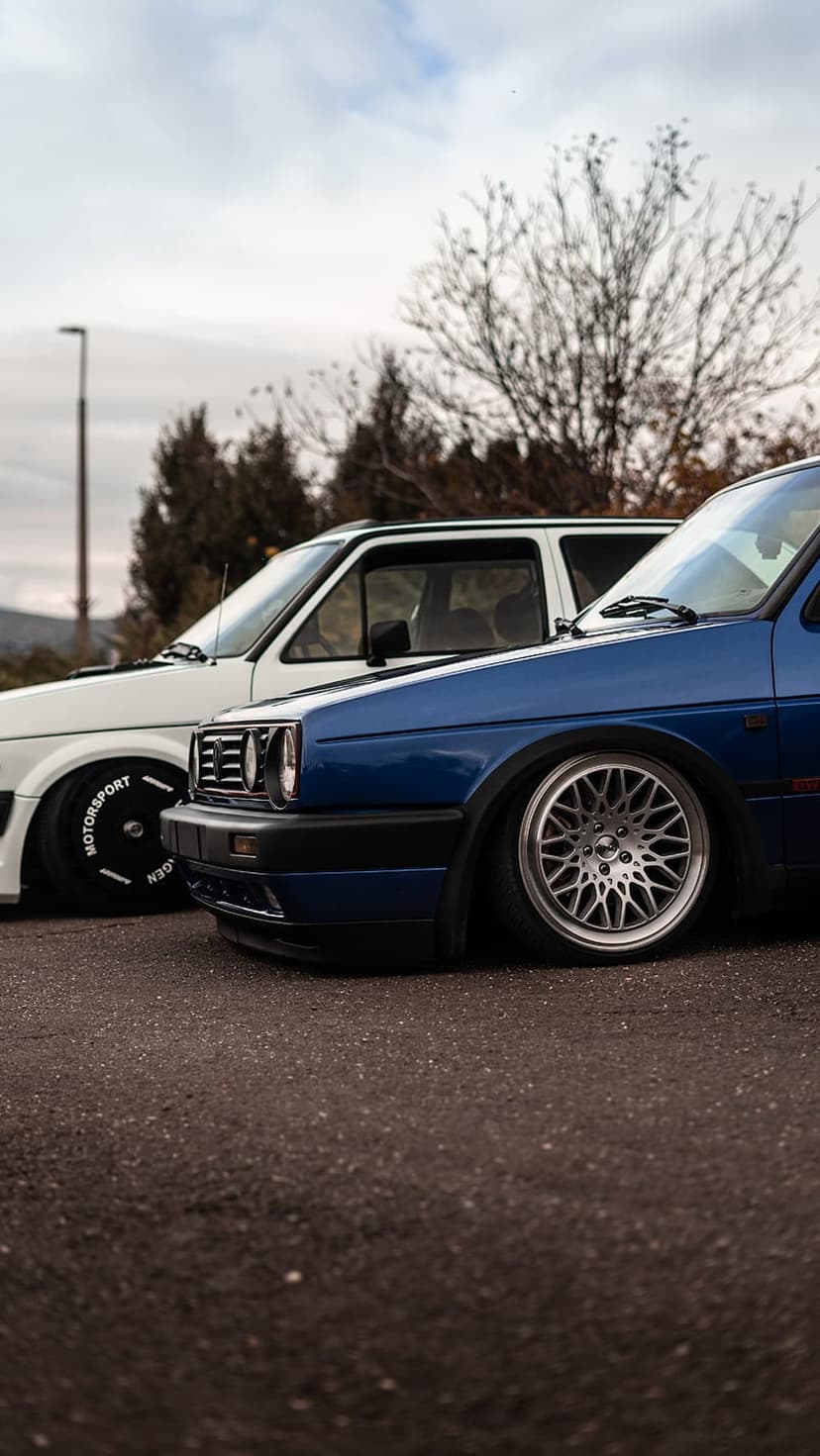 Blue and white Volkswagen Golfs with custom rims and lowered suspension parked under an overcast winter sky.