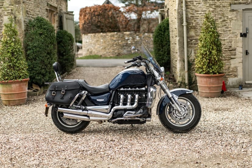 Navy blue Triumph Rocket III cruiser with chrome exhaust on a gravel path between stone walls and green bushes.