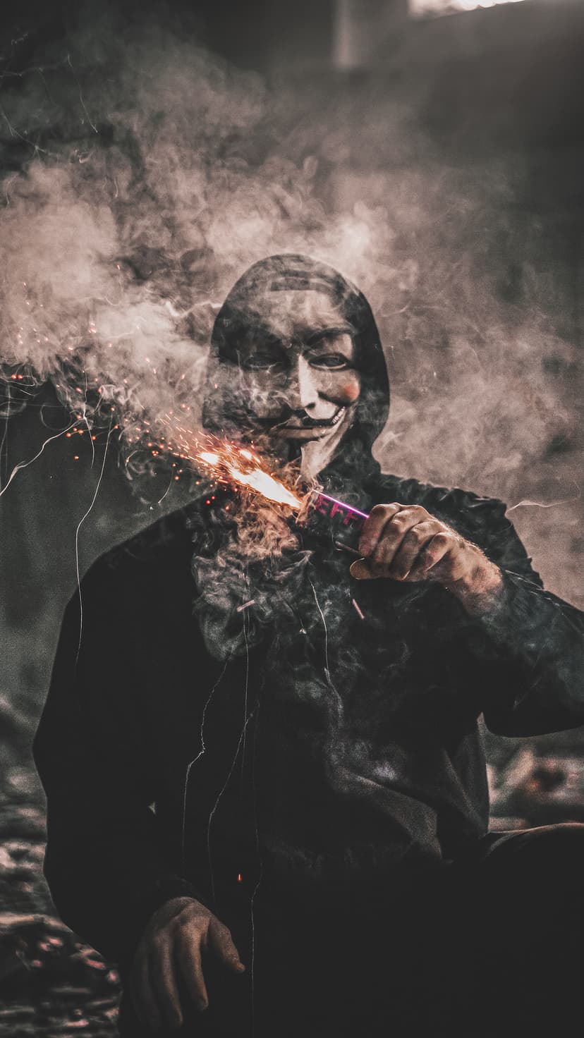 Masked figure in a black hoodie sits in a smoky urban environment with a low-angle camera perspective.