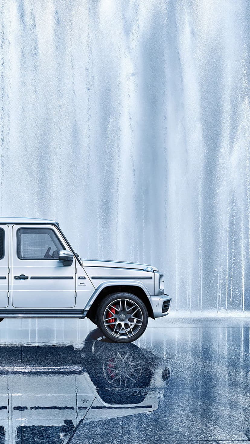 Silver Mercedes-Benz G-Wagon SUV parked near a splashing water fountain with reflections on wet ground.