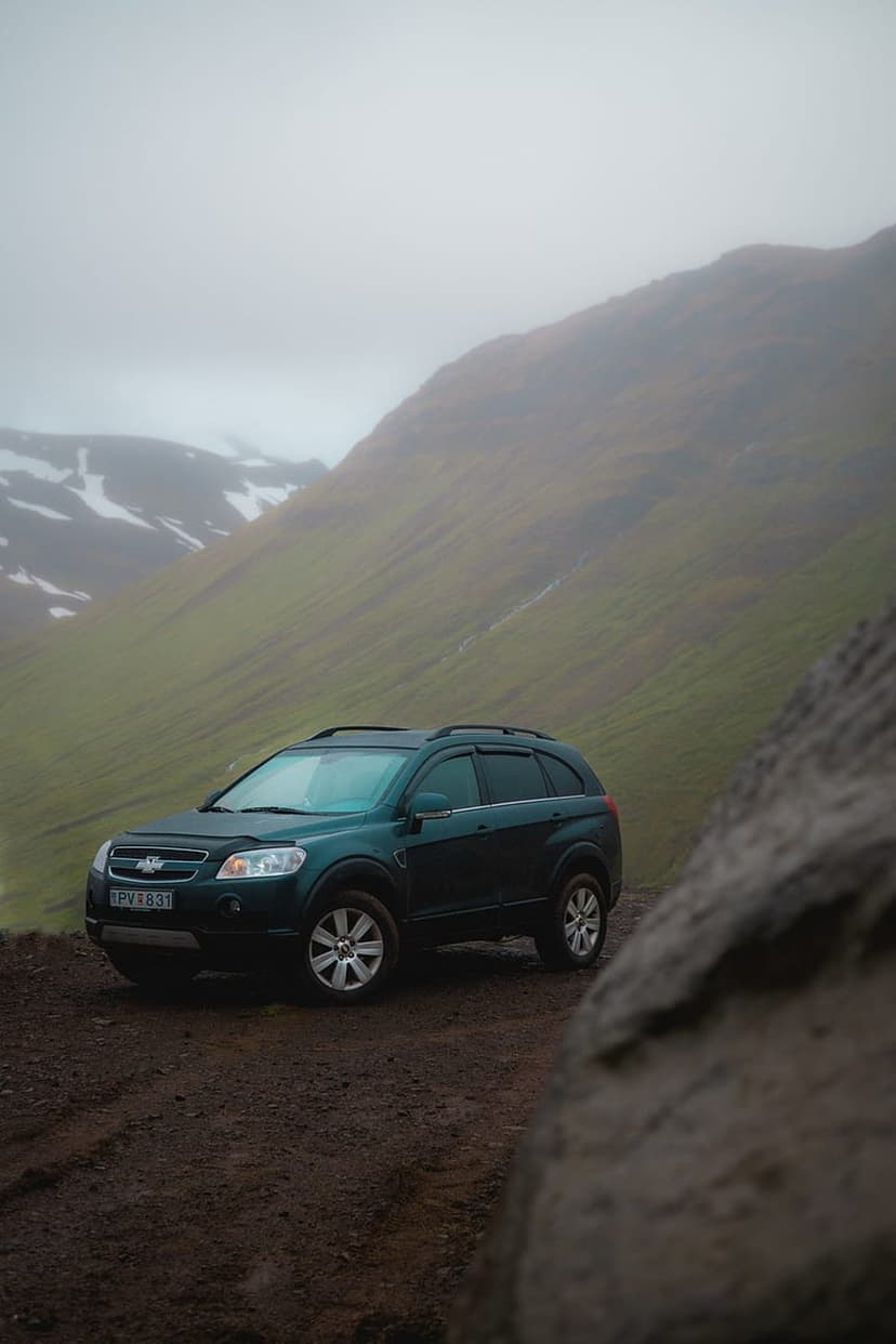 Dark green SUV parked on a gravel path near misty mountains with snow patches and a large foreground rock.