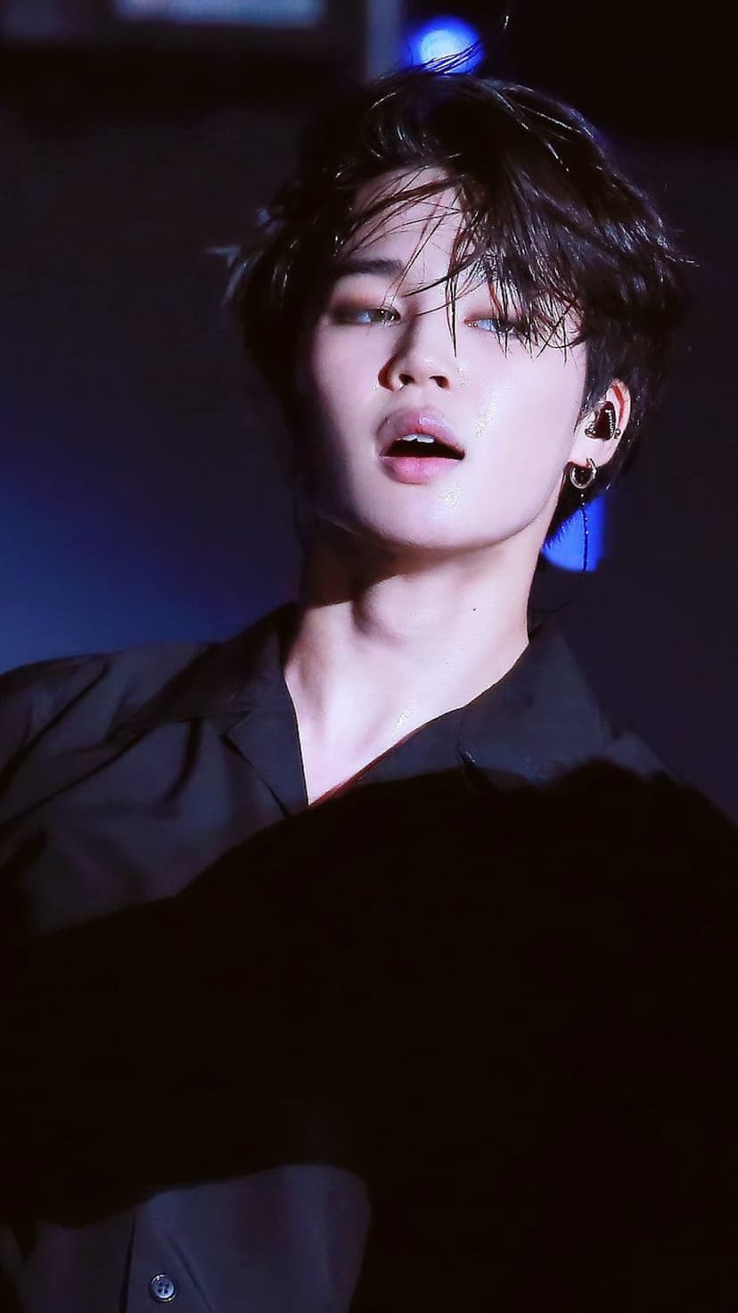 Jimin of BTS performs in a black shirt under blue spotlights with visible sweat and tousled dark hair.
