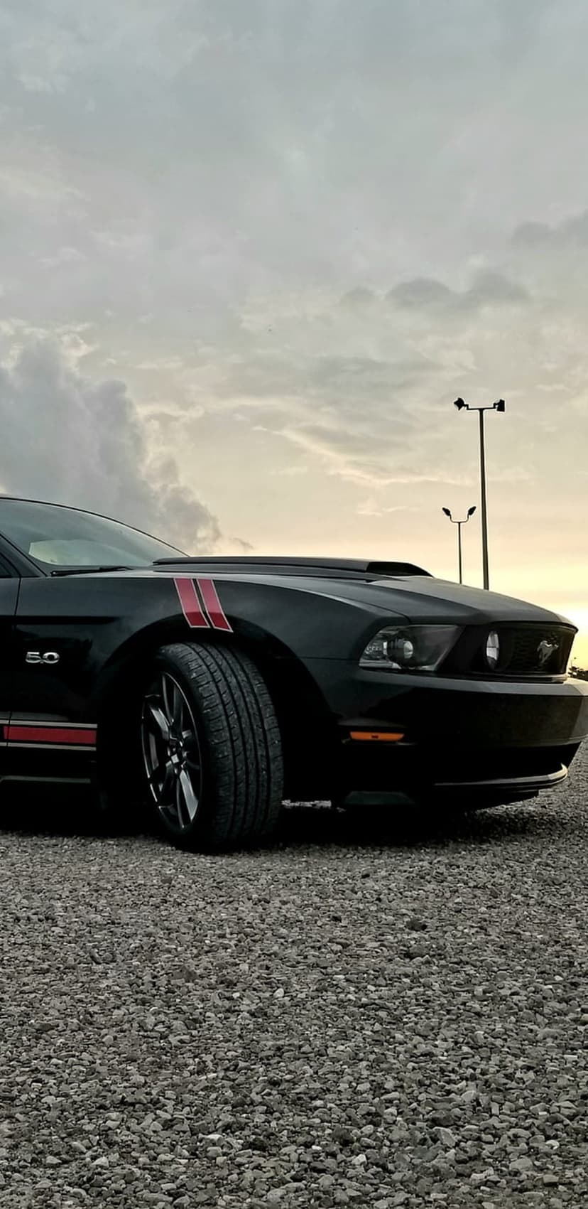 Black Mustang GT 5.0 with red stripes parked on a gravel lot under a cloudy sunset sky in a low-angle view.
