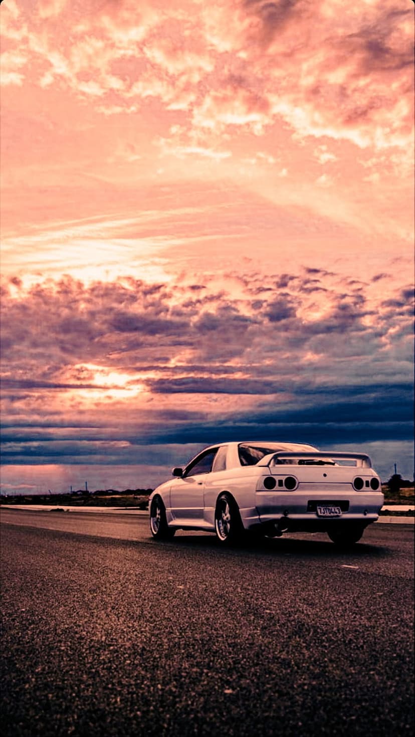 Nissan Skyline R32 GT-R with glowing red taillights parked on a road against a pink and orange sunset sky.