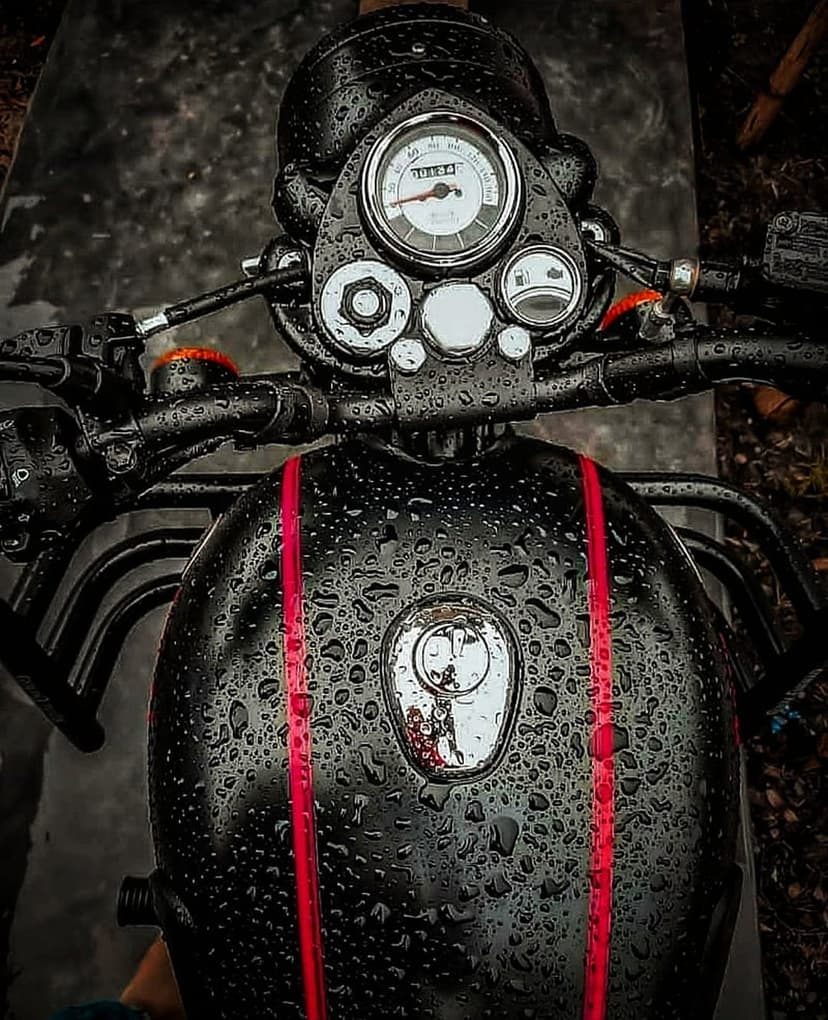 Black Royal Enfield motorcycle with water droplets on the fuel tank, red stripes, and a wet chrome instrument cluster.