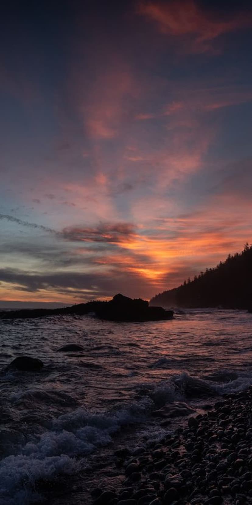 Crashing ocean waves meet a pebbled beach under a vibrant orange and pink sunset sky with silhouetted headlands.