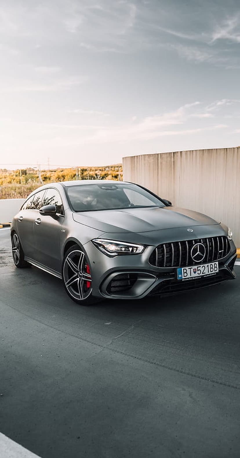 Matte gray Mercedes-AMG CLA 45 S Coupe with red calipers parked by a concrete wall under a cloudy sky.