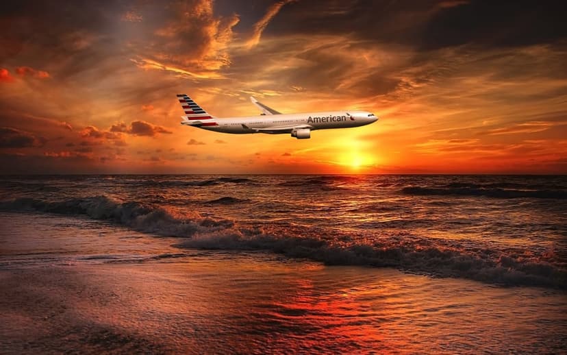 American Airlines airplane flying above ocean waves during a fiery orange and red sunset with water reflections.