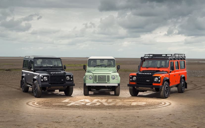Three Land Rover Defenders in black, green, and orange parked on a beach with a logo carved in the sand.