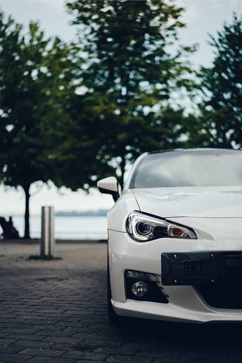 White Subaru BRZ sports car parked on a brick walkway with green trees and blue water in the background.