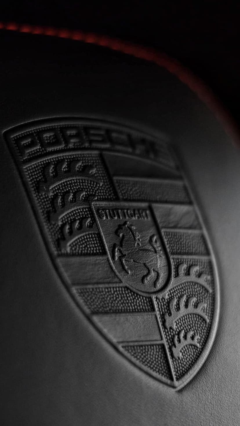 Porsche crest embossed in black on a dark pebbled surface with a single line of red stitching at the top edge.