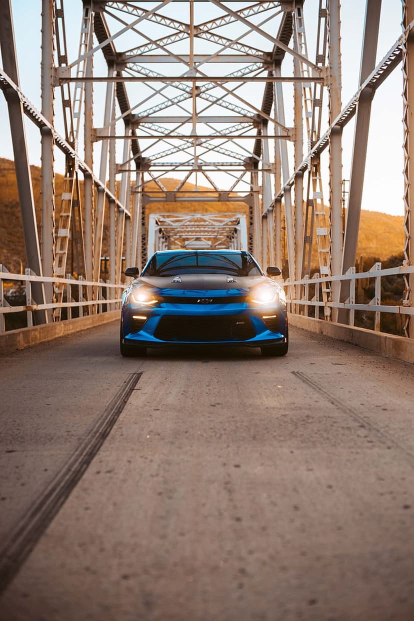 Blue Chevrolet Camaro with headlights on driving across a metal truss bridge during a golden hour sunset.