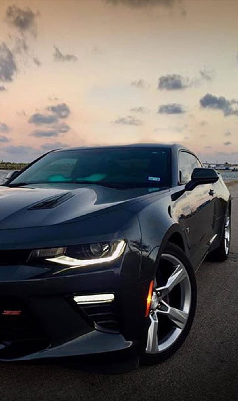 Chevrolet Camaro with lit headlights parked on a coastal road during a dramatic orange and purple sunset.