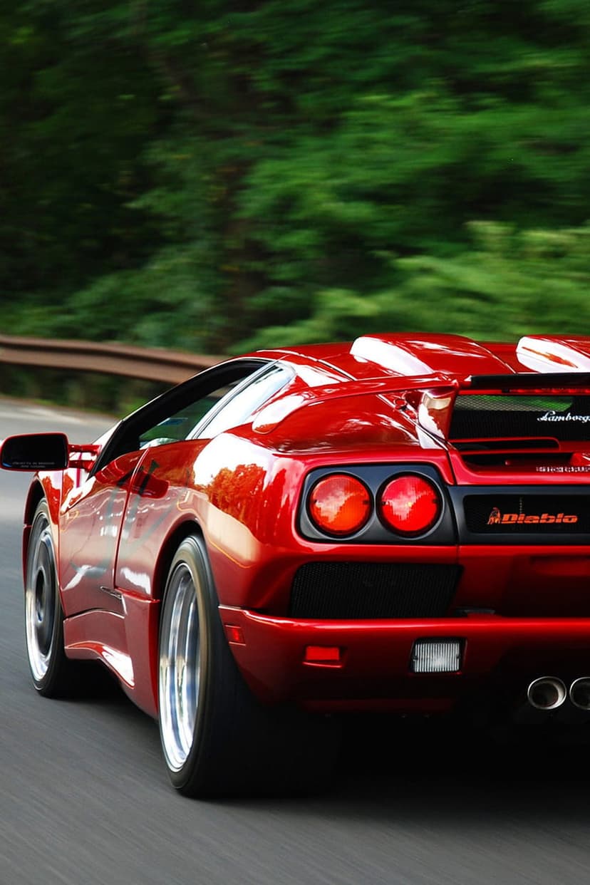 Red Lamborghini Diablo rear view with a large wing driving fast on a tree-lined road with motion blur.