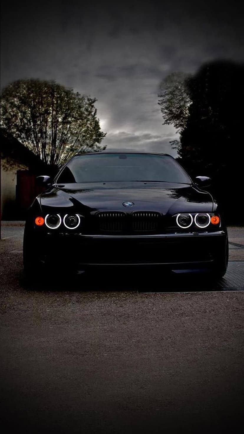 Black BMW sedan with illuminated circular headlights parked on wet pavement under a cloudy twilight sky.