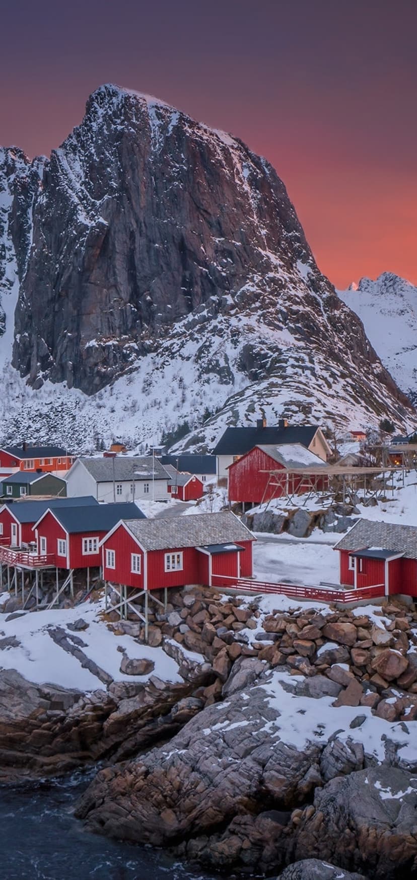Red wooden cabins sit at the foot of a snowy mountain during a vibrant orange and blue Arctic sunset.