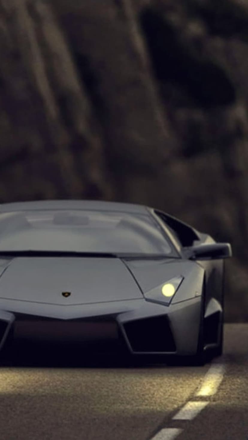 Lamborghini Aventador with glowing headlights drives on a paved road at dusk against a blurred rocky hillside.
