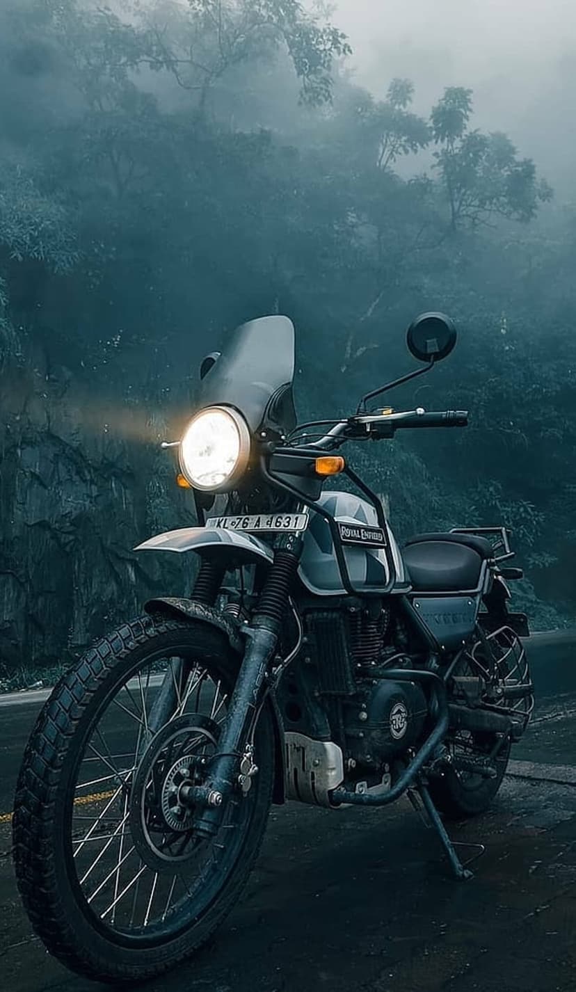 Royal Enfield Himalayan motorcycle parked on a wet road amidst dense gray mountain fog and silhouettes of pine trees.
