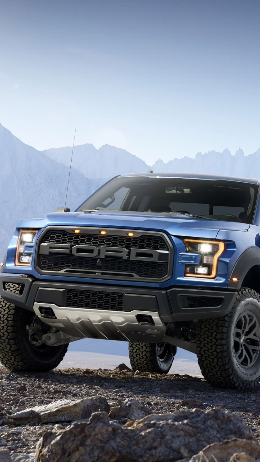 Blue Ford Raptor pickup truck parked on rocky desert ground with mountains under a clear blue sky.