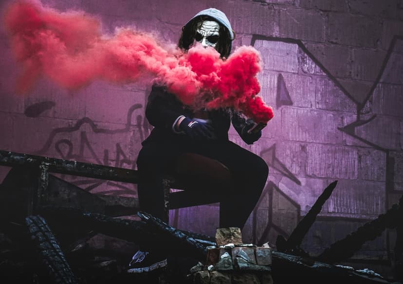 Masked person in a hooded jacket holds a venting red smoke bomb while crouching near a graffiti-covered wall.