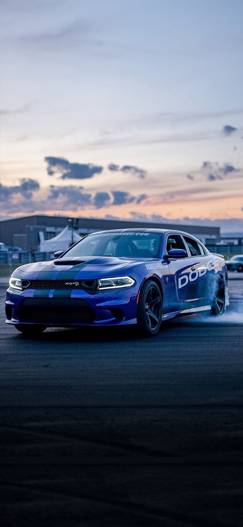 Blue Dodge Charger Hellcat drifting on asphalt with thick white tire smoke against a glowing orange sunset sky.
