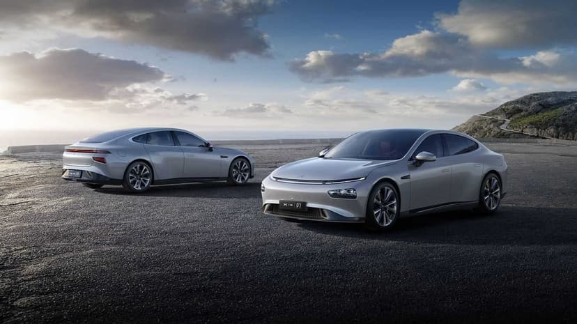 Two silver electric cars parked on a dark asphalt coastal overlook under a blue cloudy sky near mountains.