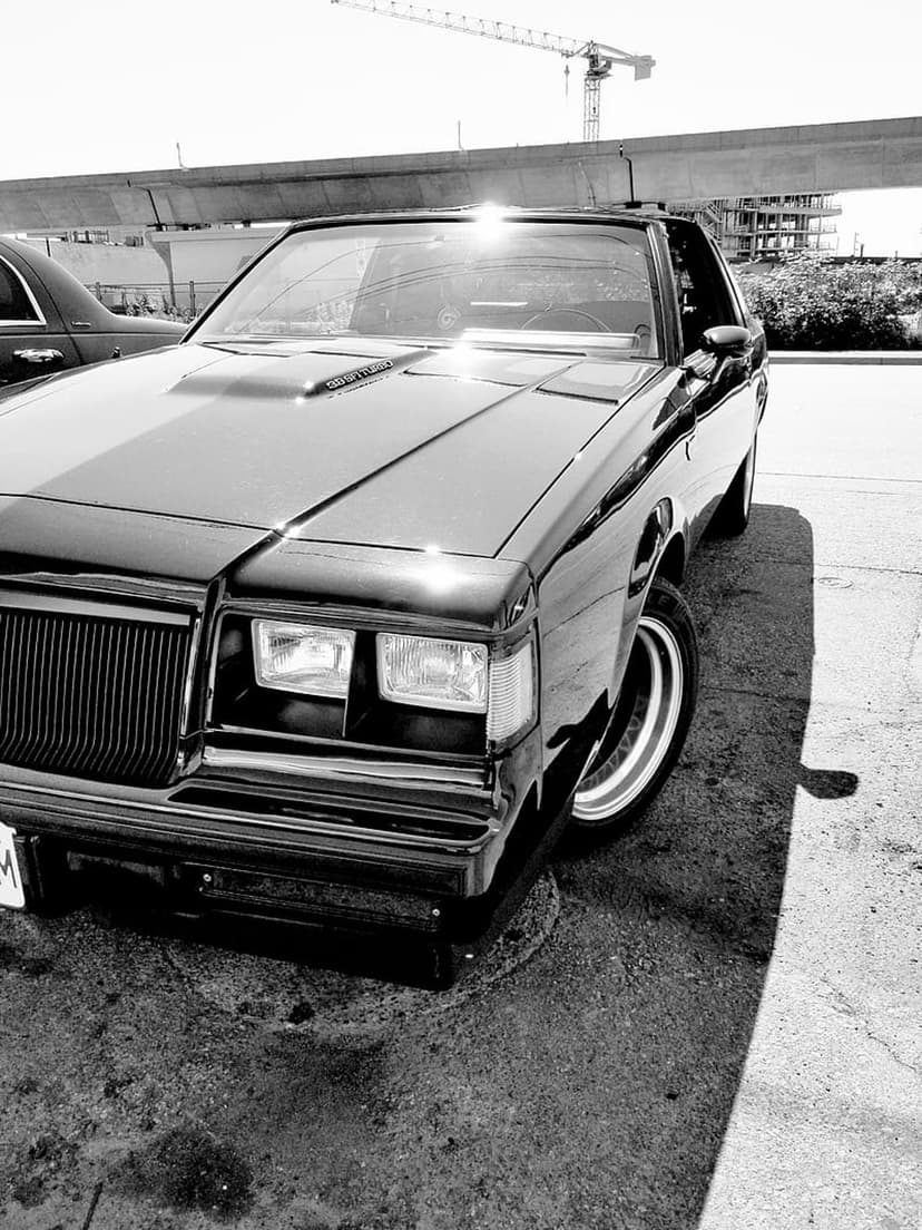 Black Buick Grand National muscle car parked on asphalt under a concrete overpass and industrial crane in sunlight.