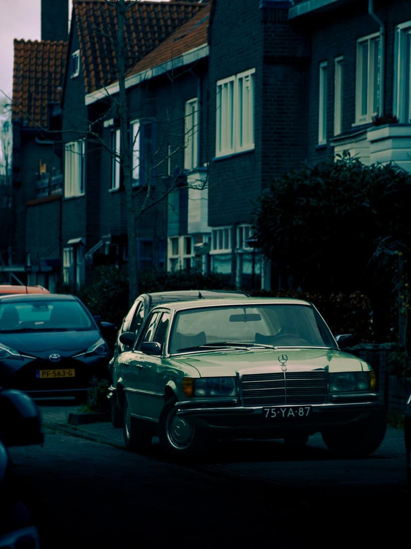 Green vintage Mercedes sedan parked in front of dark brick houses on a cloudy day with chrome accents visible.