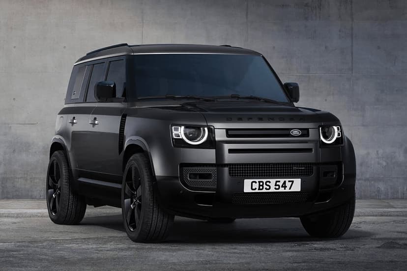 Matte black Land Rover Defender with glowing LED headlights parked against a textured gray concrete wall.