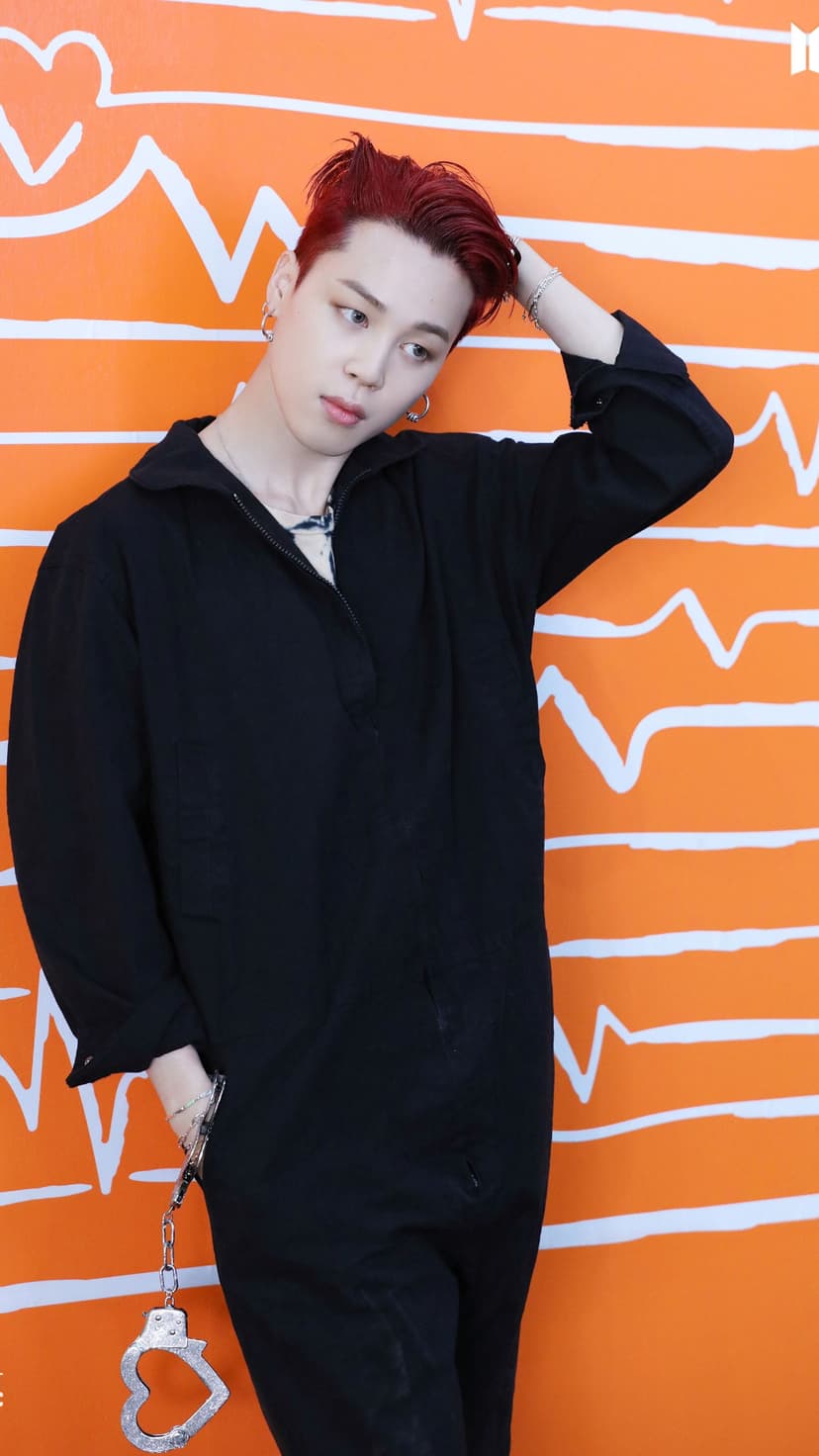 Jimin from BTS with red hair wearing a black jumpsuit and handcuffs while leaning against an orange heart wall.