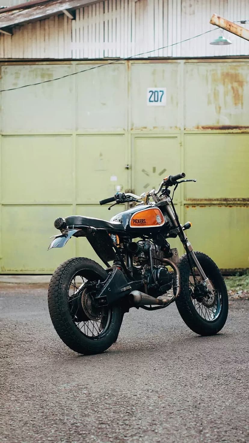 Custom black and orange scrambler motorcycle parked on asphalt in front of an olive green garage with number 207.