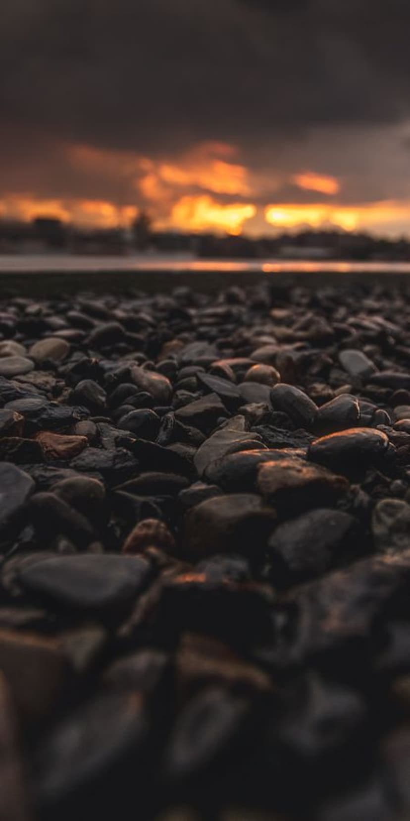 Smooth dark pebbles in the foreground lead to a bright orange sunset over a distant coastal ocean horizon.